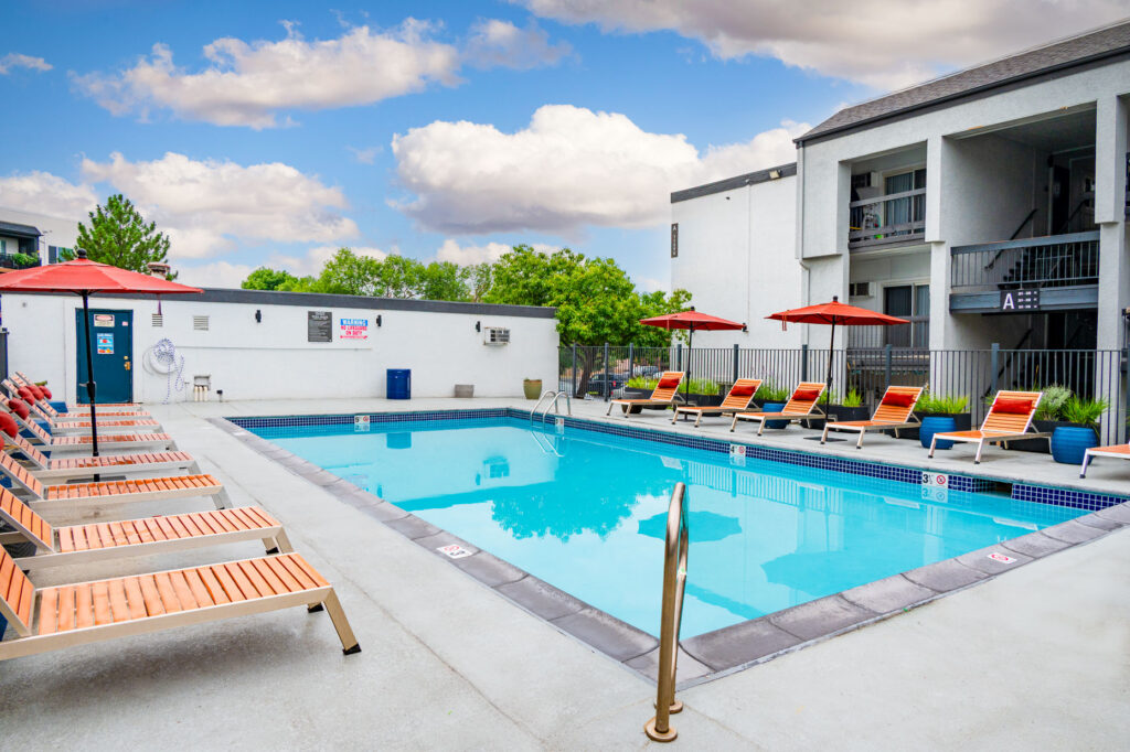 Outdoor swimming pool with clear water, surrounded by red lounge chairs and umbrellas, next to a modern white apartment building under a partly cloudy sky.