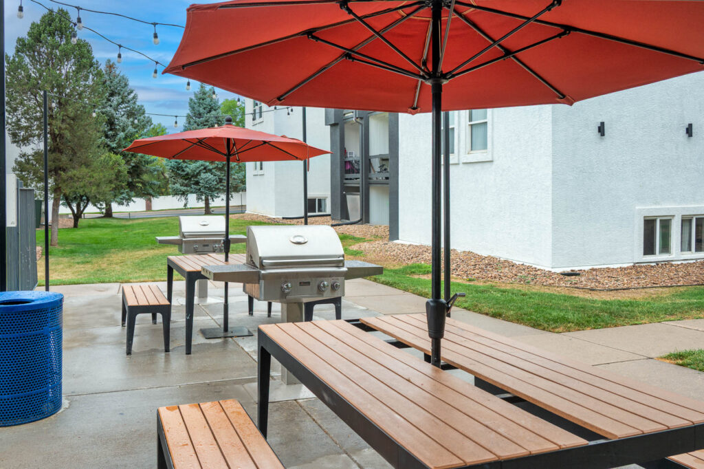 Outdoor patio with wooden picnic tables, red umbrellas, and two stainless steel grills on a concrete area. Nearby grass, trees, a blue trash can, and white apartment buildings are visible in the background.