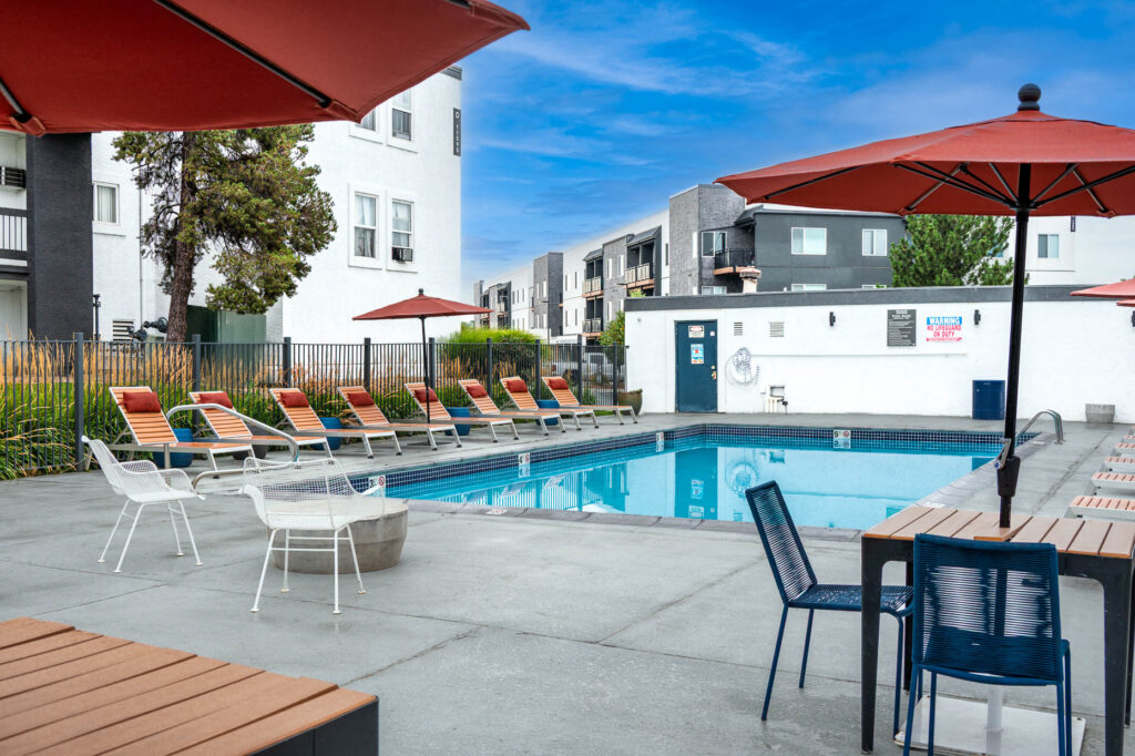 Outdoor pool area with lounge chairs, tables, and red umbrellas, surrounded by modern apartment buildings under a blue sky.